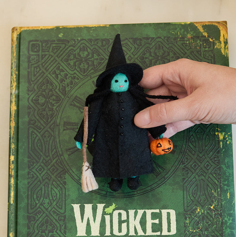 Craftspring’s Wicked Witch Trick-or-Treater ornament — with green skin, a witch’s hat, broom, and pumpkin basket — displayed on the book Wicked for a festive Halloween scene, perfect for spooky decorating and holiday gifting, ethically crafted by artisans using sustainable wool.