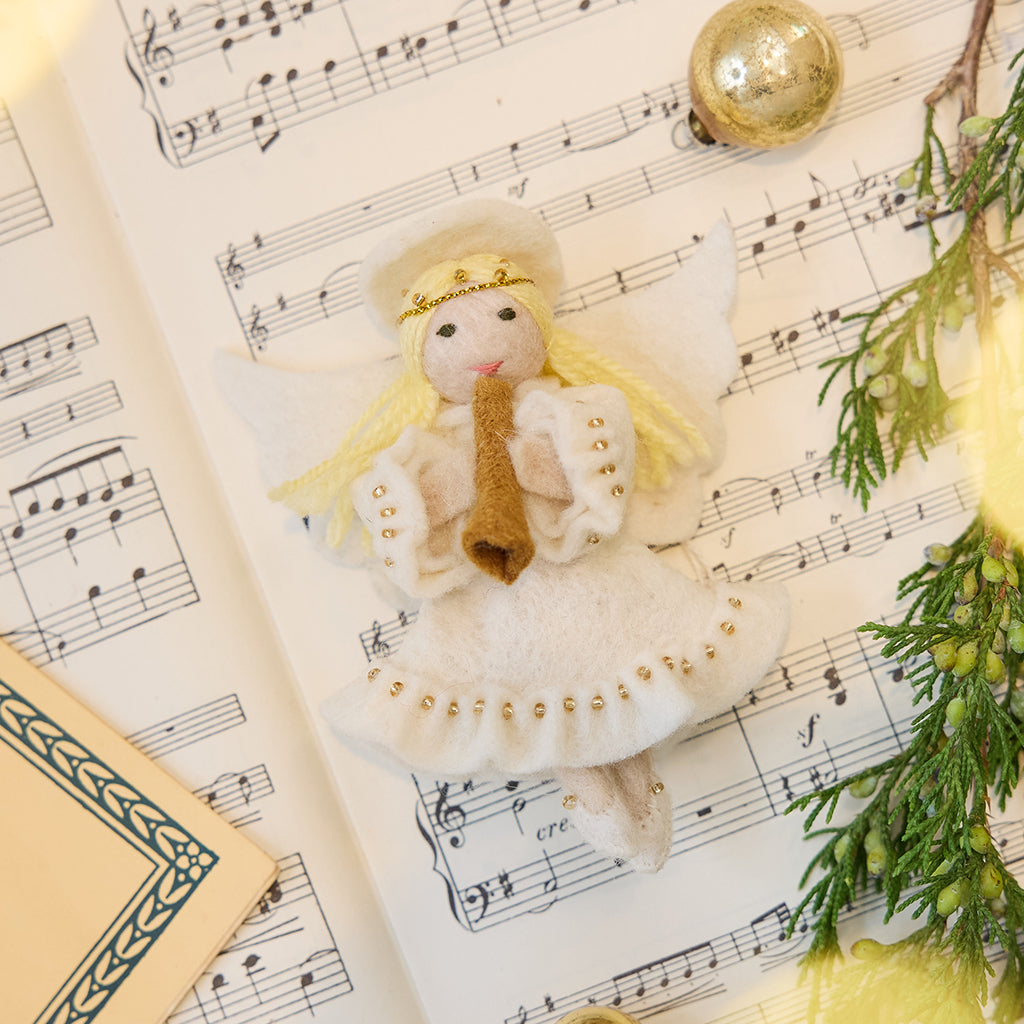 Vintage-Style Angel Playing Trumpet Ornament displayed on sheet music — handmade felt holiday ornament with intricate beaded and embroidered details, showing an angel playing a trumpet, ethically crafted by artisans using sustainable wool.