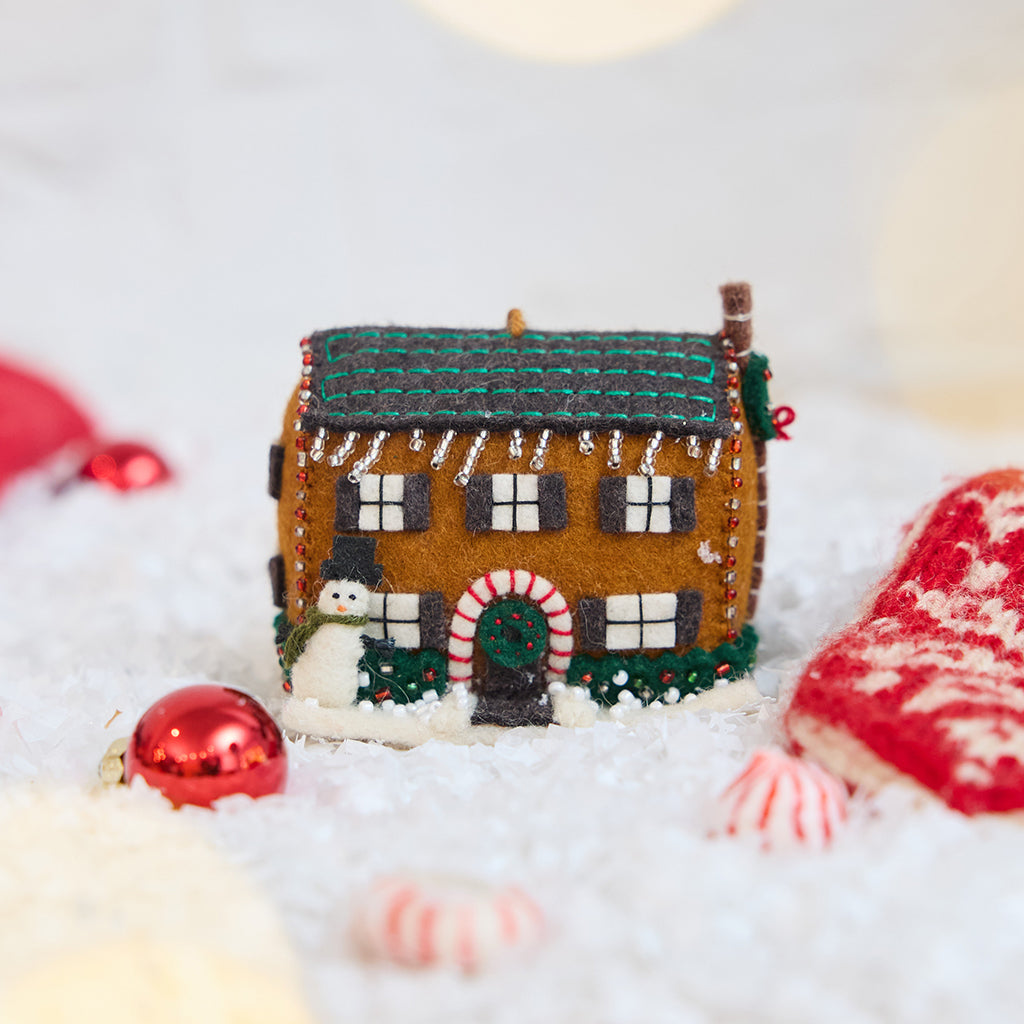 Craftspring’s Christmas House Ornament — felt house with beading, embroidery, and applique, featuring a snowman, beaded icicles, and holly bushes — displayed on snow with holiday baubles and peppermints, evoking festive warmth, playful holiday charm, and heirloom-quality gifting, ethically crafted by artisans using sustainable wool.