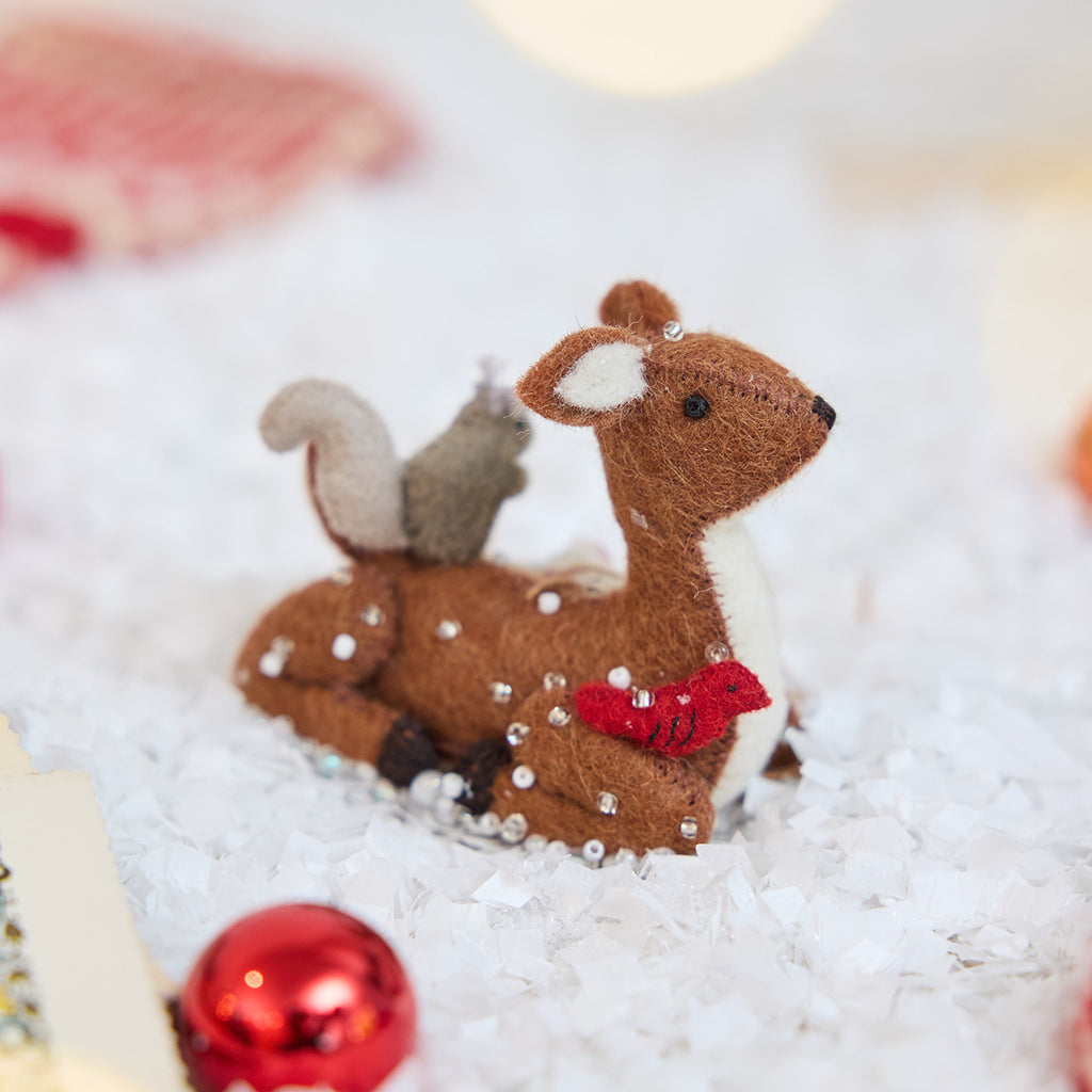 Craftspring’s Sitting Deer with Friends ornament — featuring a deer in the snow with a bunny, squirrel, and bird nestled around it, with intricate beaded snow and embroidered details — displayed in the snow with bobbles for a festive Christmas scene, ethically crafted by artisans using sustainable wool.