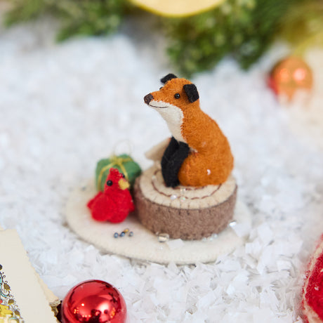 Craftspring’s Fox on Stump ornament — a fox perched on a stump with a green present and red cardinal — displayed on snow with bobbles and evergreens for a cozy woodland Christmas scene, perfect for holiday gifting, ethically crafted by artisans using sustainable wool.