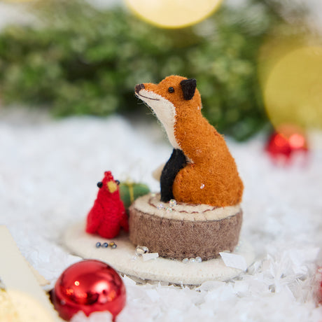 Craftspring’s Fox on Stump ornament — a fox perched on a stump with a green present and red cardinal — displayed on snow with bobbles and evergreens for a cozy woodland Christmas scene, perfect for holiday gifting, ethically crafted by artisans using sustainable wool.