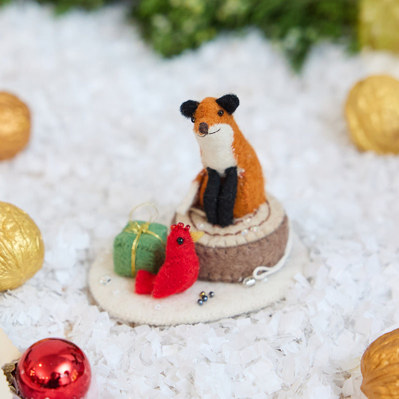 Craftspring’s Fox on Stump ornament — a fox perched on a stump with a green present and red cardinal — displayed on snow with bobbles and evergreens for a cozy woodland Christmas scene, perfect for holiday gifting, ethically crafted by artisans using sustainable wool.