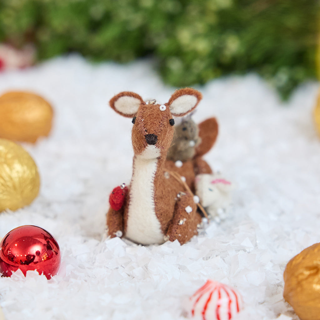 Craftspring’s Sitting Deer with Friends ornament — featuring a deer in the snow with a bunny, squirrel, and bird nestled around it, with intricate beaded snow and embroidered details — displayed in the snow with bobbles for a festive Christmas scene, ethically crafted by artisans using sustainable wool.