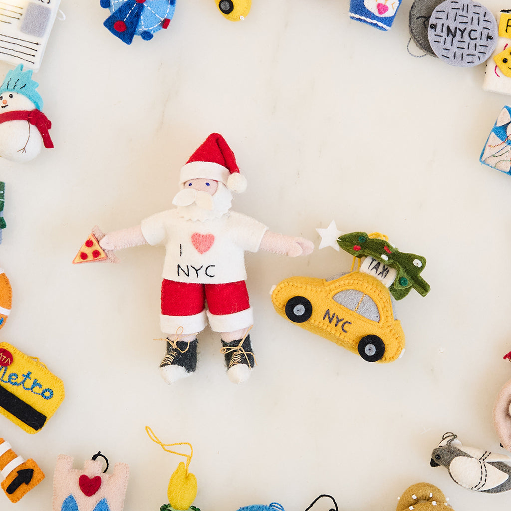Craftspring’s Christmas in the City NYC Ornaments Bundle — includes NYC Santa Ornament and Holiday Taxi Ornament — displayed together in a cozy holiday setting, highlighting playful city-themed designs with NYC Santa holding a pizza slice and the taxi with a Christmas tree on top, evoking festive warmth and heirloom-quality Christmas gifting, ethically crafted by artisans using sustainable wool.
