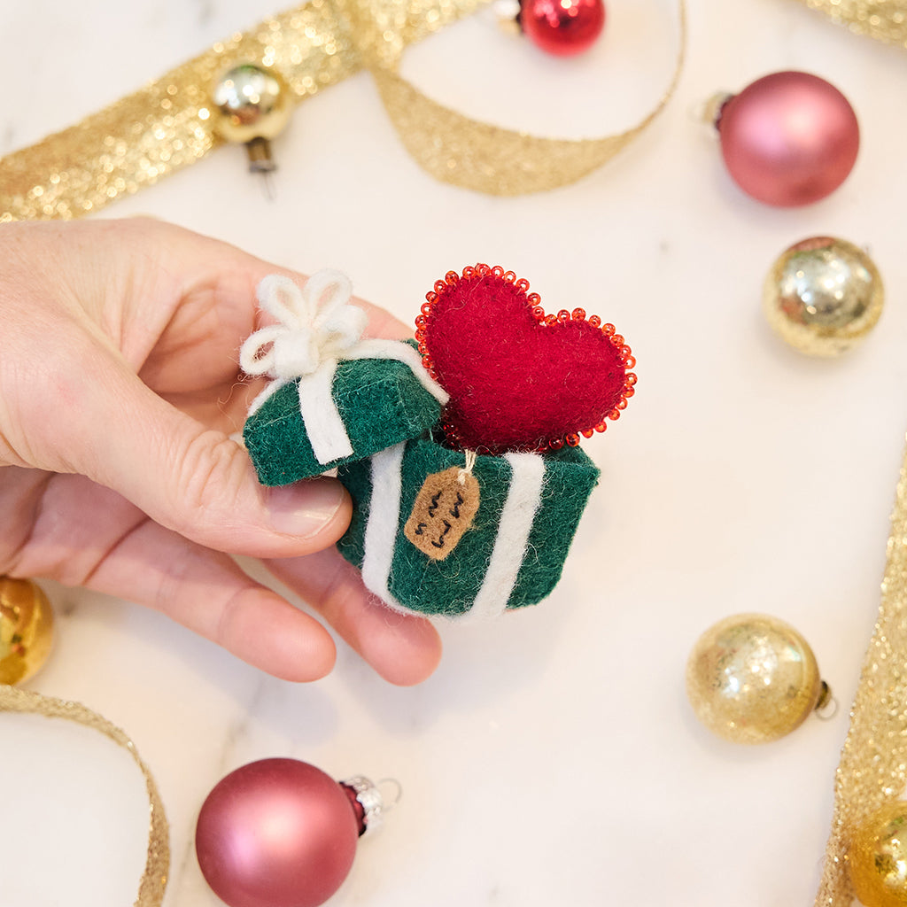 Craftspring’s Gift from the Heart Ornament — felt heart ornament with beaded and embroidered details including a gift tag — held in hand over Christmas bobbles and ribbons, creating a warm and festive holiday scene, perfect for gifting, ethically crafted by artisans using sustainable wool.