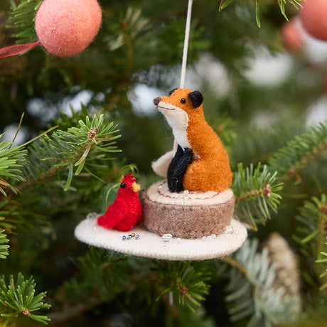 Craftspring’s Fox on Stump ornament — featuring a fox perched on a stump with a green present and red cardinal at its side — displayed on a Christmas tree for a festive woodland holiday scene, perfect for gifting, ethically crafted by artisans using sustainable wool.