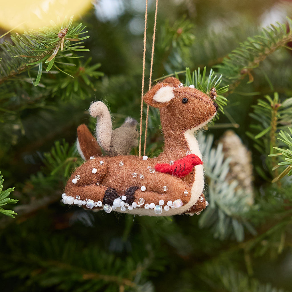 Craftspring’s Sitting Deer with Friends ornament — featuring a deer with a bunny, squirrel, and bird nestled around it, with intricate beaded snow and embroidered details — displayed on a Christmas tree for a festive woodland holiday scene, ethically crafted by artisans using sustainable wool.