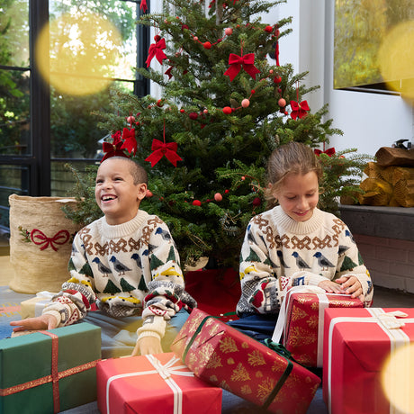 Craftspring’s NYC Holiday 100% Alpaca Sweater — heirloom-quality crewneck knit from 100% sustainably harvested baby alpaca wool, featuring intricate NYC jacquard motifs including bagels, taxis, and pigeons — ethically crafted by artisans in Peru. Two kids wear the sweaters while opening presents under the Christmas tree, capturing holiday joy and gifting warmth.