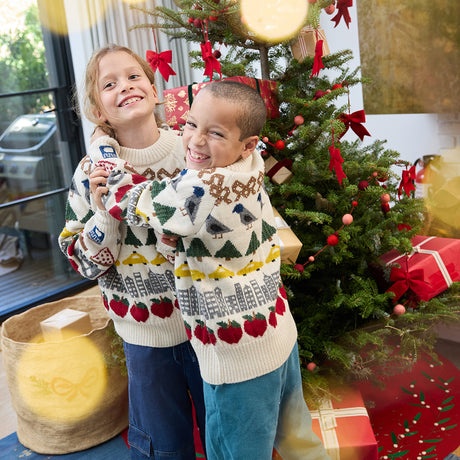 Craftspring’s NYC Holiday 100% Alpaca Sweater — soft crewneck of sustainably harvested baby alpaca wool, adorned with intricate jacquard NYC icons such as bagels, taxi cabs, and pigeons — heirloom-quality holiday knitwear, ethically crafted by artisans in Peru. Two kids wear the sweaters in front of a Christmas tree, radiating festive cheer and Christmas gifting spirit.