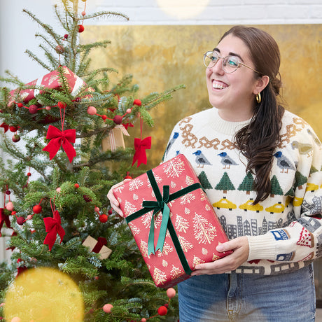 Craftspring’s NYC Holiday 100% Alpaca Sweater — heirloom-quality crewneck crafted of ultra-soft, sustainably harvested baby alpaca wool with intricate jacquard NYC motifs such as bagels, pigeons, and taxi cabs — ethically made by artisans in Peru. A woman wears the sweater in front of a Christmas tree, radiating Christmas elegance, warmth, and holiday gifting joy.