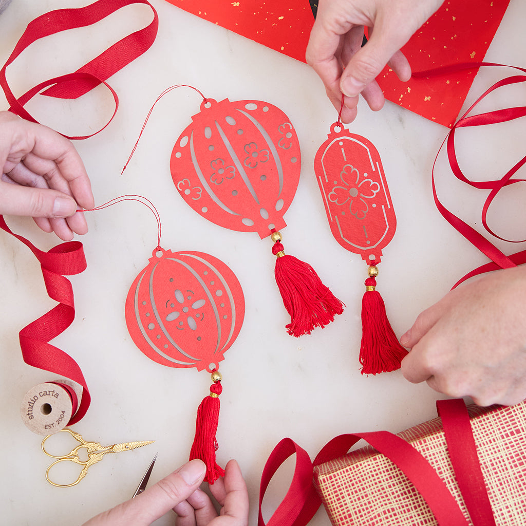 Paper Lantern Ornaments held in hand among loops of red ribbon, glowing with delicate cutout designs. Ethically handmade by artisans for Lunar New Year gifting and timeless decor.