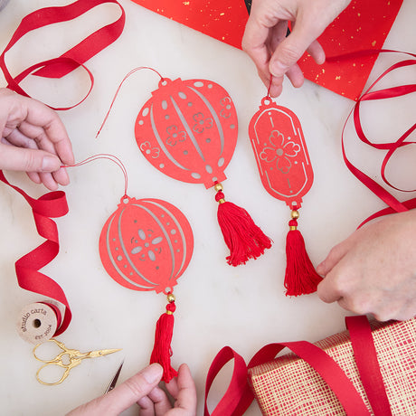 Paper Lantern Ornaments held in hand among loops of red ribbon, glowing with delicate cutout designs. Ethically handmade by artisans for Lunar New Year gifting and timeless decor.