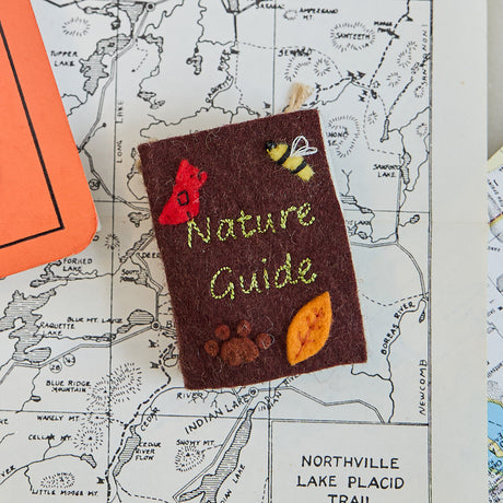 Miniature hand-sewn felt book ornament titled Nature Guide, featuring embroidered and appliquéd illustrations of plants, animals, and woodland scenes. Made from merino-blend wool with whimsical details and a hanging string. Measures approximately 2.5 inches tall by 3 inches wide. A charming tribute to the outdoors and nature lovers.