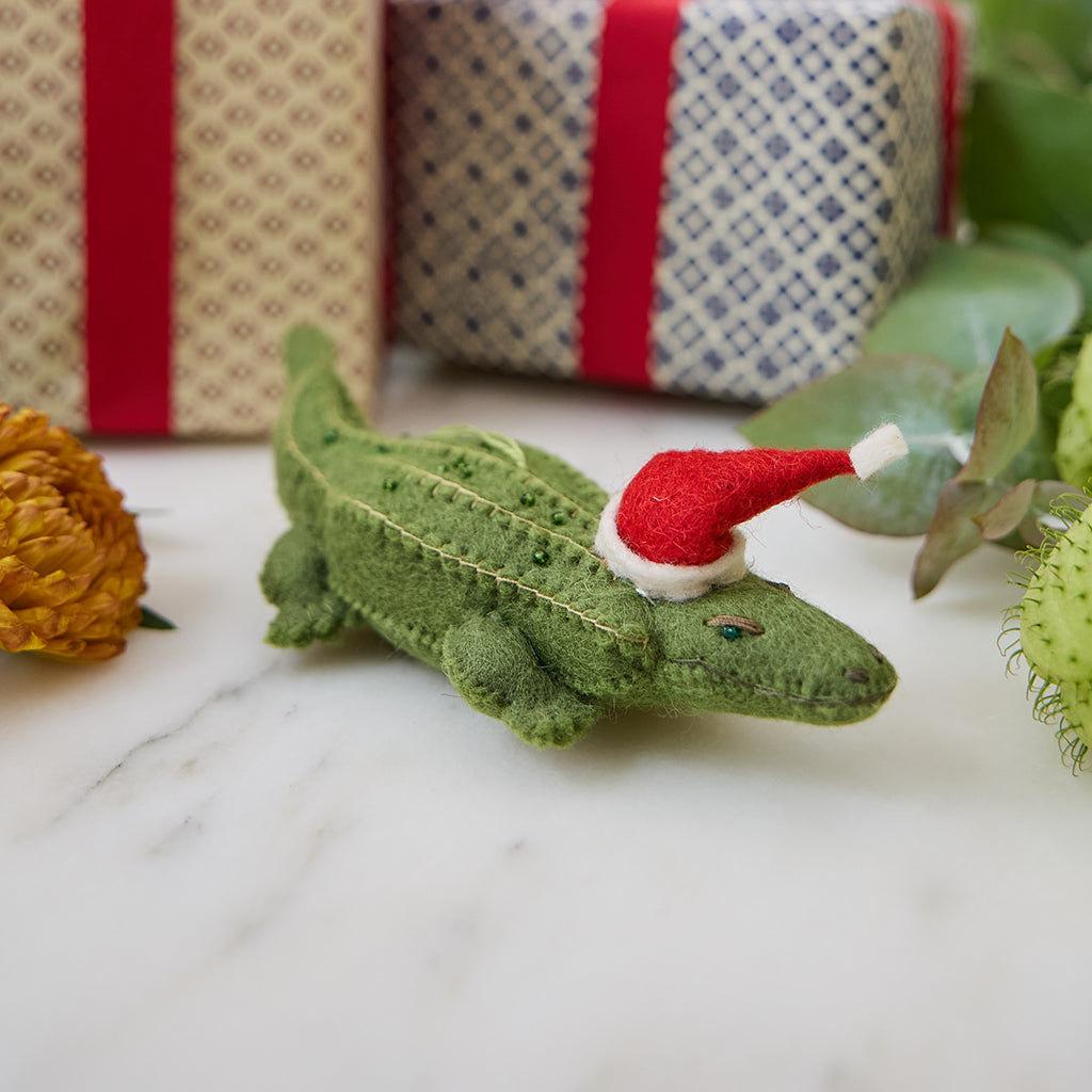 Craftspring’s Santa Alligator Ornament — felt alligator with a beautiful face and embroidered and beaded back ridges, displayed among presents for a festive holiday scene — heirloom-quality Christmas ornament, perfect for gifting, ethically crafted by artisans using sustainable wool.