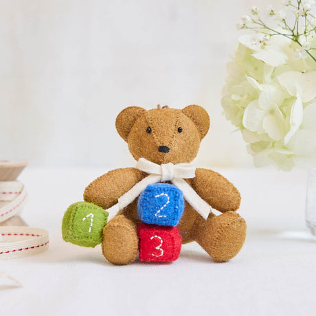 Classic and comforting, this adorable teddy bear ornament is crafted from soft, natural wool felt and finished with a gentle cream scarf. Nestled beside colorful number blocks, it captures the sweetness of early childhood and the joy of new beginnings.