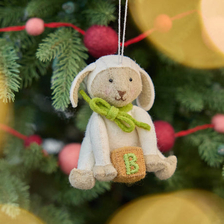 Hanging on a soft glowing Christmas tree. Soft, sweet, and timeless, this charming lamb ornament is carefully handcrafted from natural wool felt. With its floppy ears, gentle expression, and bright green bow, it holds a tiny embroidered “B” block—symbolizing baby’s earliest milestones.&nbsp;