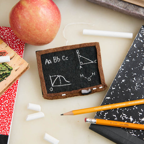 Miniature chalkboard with educational drawings, pencils, and an apple on a light surface. 
A double-sided handmade felt school blackboard ornament featuring a world map on one side and classic math formulas on the other. An ode to old-school learning and the teachers that inspired us.