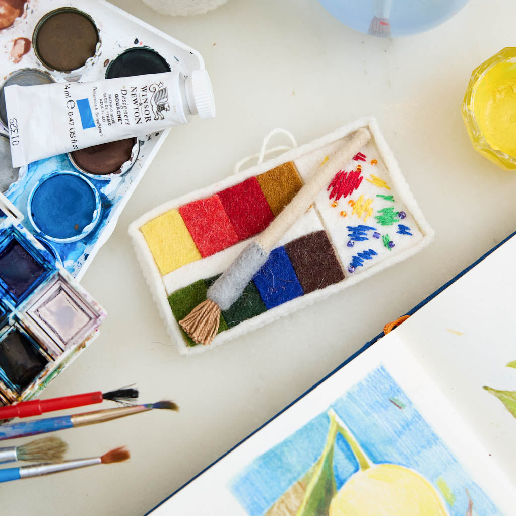 A small celebration of color in miniature form, our Paint Palette Ornament captures the familiar beauty of an artist’s workspace. Soft blocks of felted color sit alongside a tiny paintbrush with splashing embroidered brushstrokes, as if paused mid-creation.