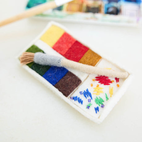 A small celebration of color in miniature form, our Paint Palette Ornament captures the familiar beauty of an artist’s workspace. Soft blocks of felted color sit alongside a tiny paintbrush with splashing embroidered brushstrokes, as if paused mid-creation.
