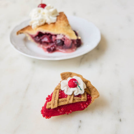 A true slice of nostalgia, our Cherry Pie Slice Ornament captures the timeless charm of one of America's favorite desserts. Handcrafted from natural wool felt, this delightful ornament features a rich cherry filling accented with sparkling red beadwork, a beautifully detailed lattice crust, a dollop of whipped cream, and a bright cherry on top.
