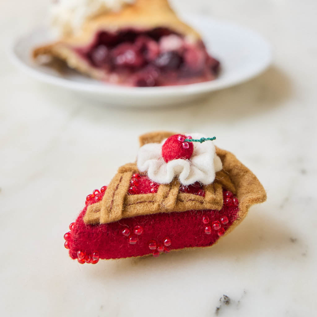 A true slice of nostalgia, our Cherry Pie Slice Ornament captures the timeless charm of one of America's favorite desserts. Handcrafted from natural wool felt, this delightful ornament features a rich cherry filling accented with sparkling red beadwork, a beautifully detailed lattice crust, a dollop of whipped cream, and a bright cherry on top.