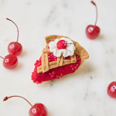 A true slice of nostalgia, our Cherry Pie Slice Ornament captures the timeless charm of one of America's favorite desserts. Handcrafted from natural wool felt, this delightful ornament features a rich cherry filling accented with sparkling red beadwork, a beautifully detailed lattice crust, a dollop of whipped cream, and a bright cherry on top.