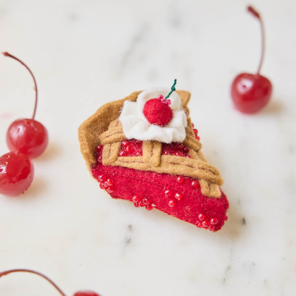 A true slice of nostalgia, our Cherry Pie Slice Ornament captures the timeless charm of one of America's favorite desserts. Handcrafted from natural wool felt, this delightful ornament features a rich cherry filling accented with sparkling red beadwork, a beautifully detailed lattice crust, a dollop of whipped cream, and a bright cherry on top.