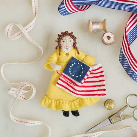 Celebrate the legendary maker associated with America’s first flag with our handcrafted Betsy Ross Ornament. Dressed in a bright colonial gown and holding the iconic early American flag, Betsy honors the spirit of independence, ingenuity, and handmade tradition.