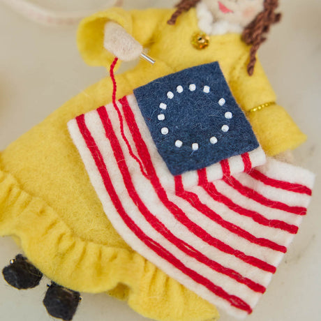Celebrate the legendary maker associated with America’s first flag with our handcrafted Betsy Ross Ornament. Dressed in a bright colonial gown and holding the iconic early American flag, Betsy honors the spirit of independence, ingenuity, and handmade tradition.