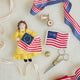Celebrate the legendary maker associated with America’s first flag with our handcrafted Betsy Ross Ornament. Dressed in a bright colonial gown and holding the iconic early American flag, Betsy honors the spirit of independence, ingenuity, and handmade tradition.