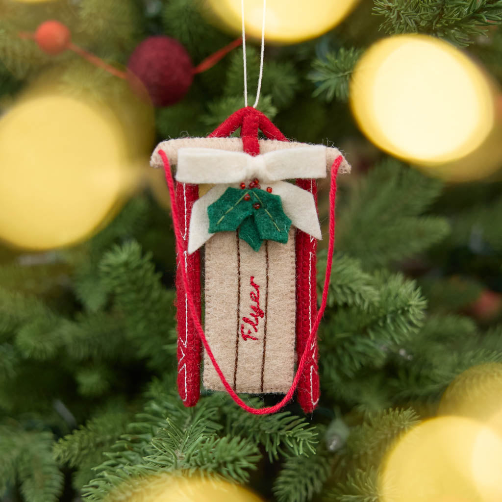 Our Red Flyer Sled Ornament captures the nostalgia of childhood winters and treasured holiday traditions. Inspired by classic wooden sleds, this charming keepsake features bright red runners, a cozy neutral-toned seat embroidered with "Flyer," and a festive holly sprig nestled beneath a soft white bow. 