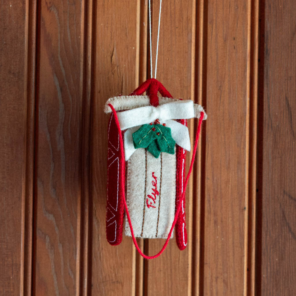Rosebud! Our Red Flyer Sled Ornament captures the nostalgia of childhood winters and treasured holiday traditions. Inspired by classic wooden sleds, this charming keepsake features bright red runners, a cozy neutral-toned seat embroidered with "Flyer," and a festive holly sprig nestled beneath a soft white bow.