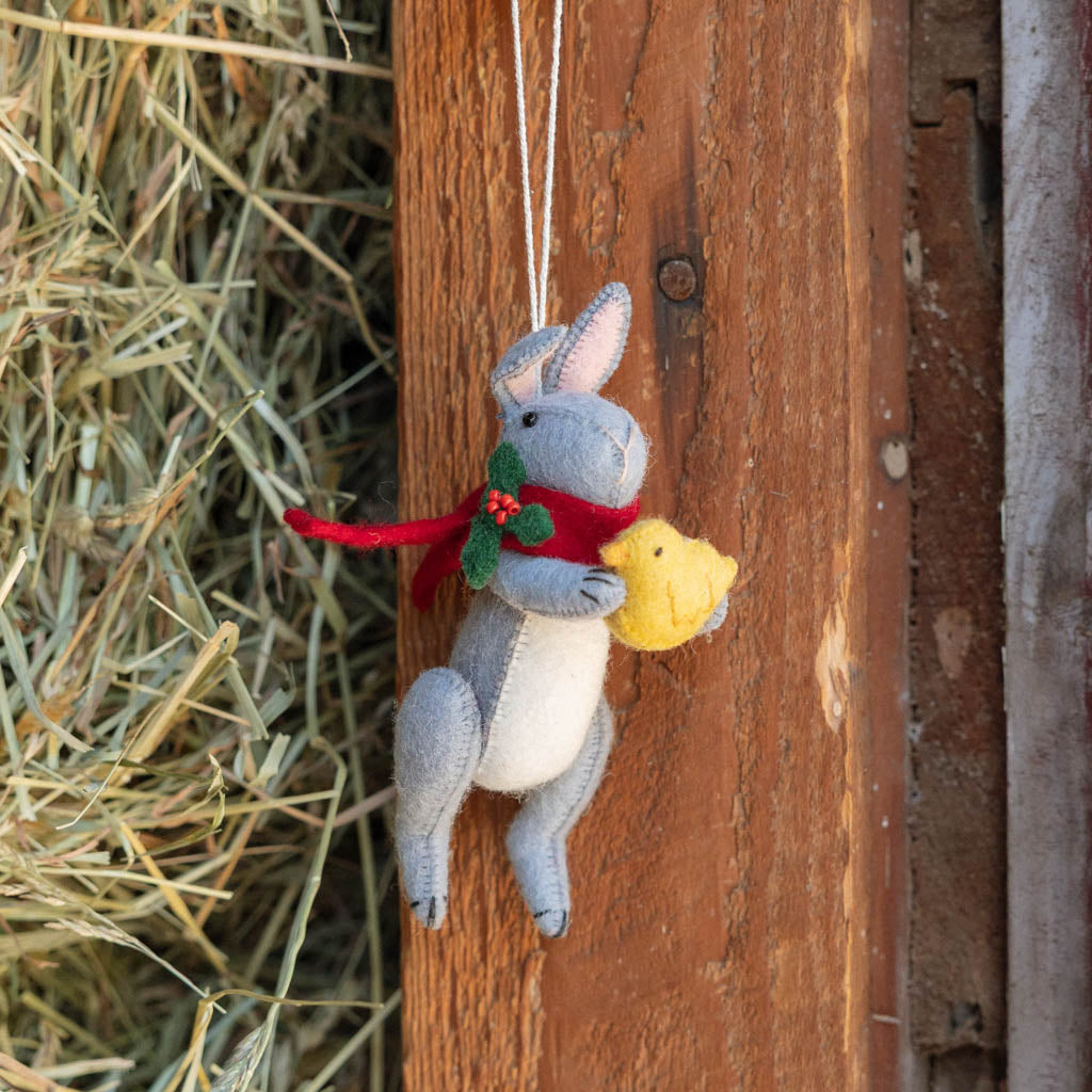 Our Gray Holiday Bunny with Chick Ornament captures a heartwarming moment of gentle friendship. Wrapped in a cozy red scarf adorned with festive holly leaves and tiny red berries, this sweet gray bunny lovingly cradles a fluffy yellow chick, creating a scene that's full of warmth, comfort, and Christmas cheer.