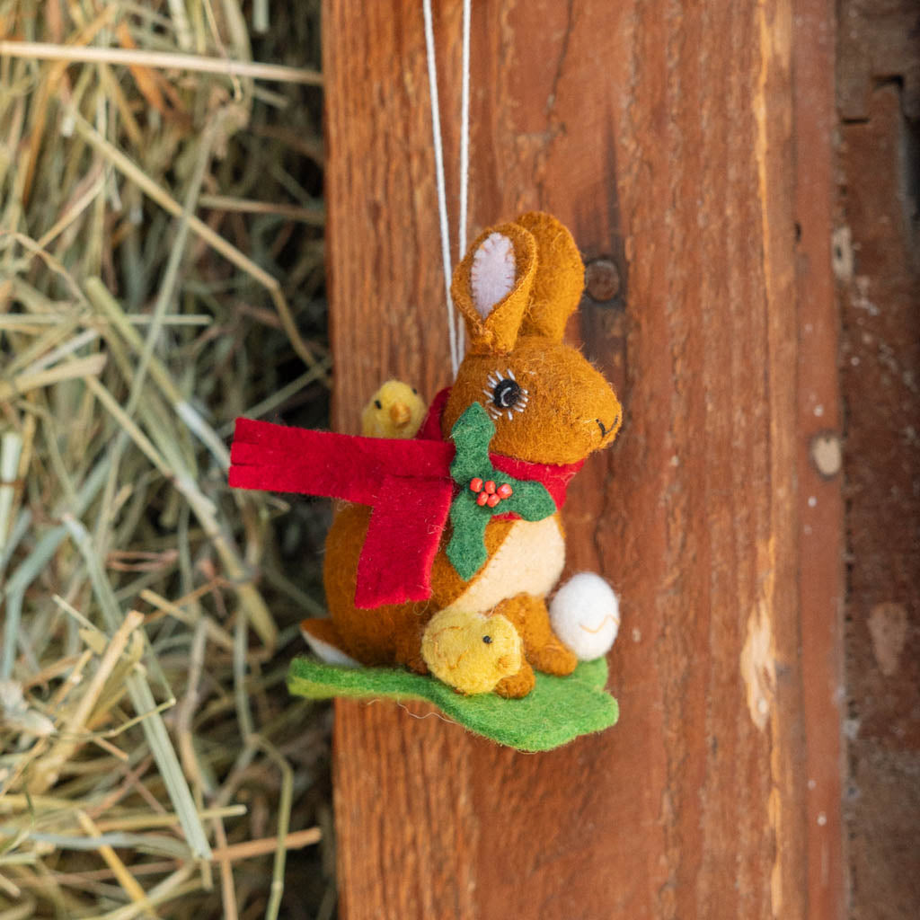 Our Brown Holiday Bunny with Chick Ornament is a sweet story of kindness and holiday cheer. Wrapped in a cozy crimson scarf adorned with felt holly and tiny red beads, this gentle brown bunny sits besides fluffy yellow chicks and a freshly hatched egg, capturing the magic of winter and the simple joy of being together.