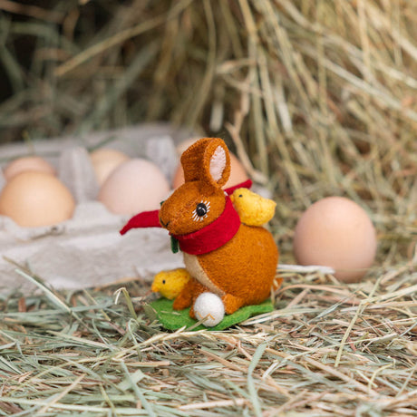 Our Brown Holiday Bunny with Chick Ornament is a sweet story of kindness and holiday cheer. Wrapped in a cozy crimson scarf adorned with felt holly and tiny red beads, this gentle brown bunny sits besides fluffy yellow chicks and a freshly hatched egg, capturing the magic of winter and the simple joy of being together.