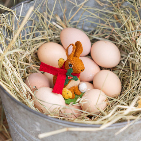 Our Brown Holiday Bunny with Chick Ornament is a sweet story of kindness and holiday cheer. Wrapped in a cozy crimson scarf adorned with felt holly and tiny red beads, this gentle brown bunny sits besides fluffy yellow chicks and a freshly hatched egg, capturing the magic of winter and the simple joy of being together.
