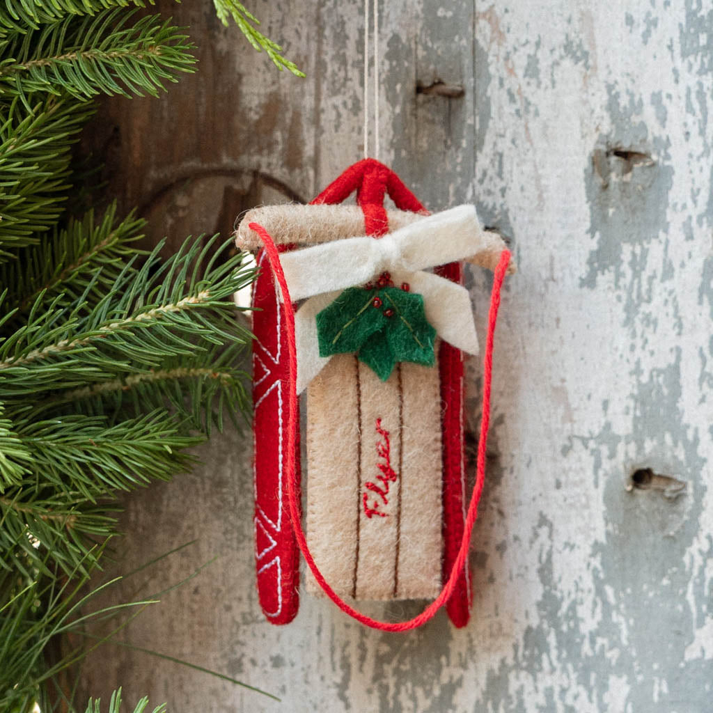 Rosebud! Our Red Flyer Sled Ornament captures the nostalgia of childhood winters and treasured holiday traditions. Inspired by classic wooden sleds, this charming keepsake features bright red runners, a cozy neutral-toned seat embroidered with "Flyer," and a festive holly sprig nestled beneath a soft white bow.