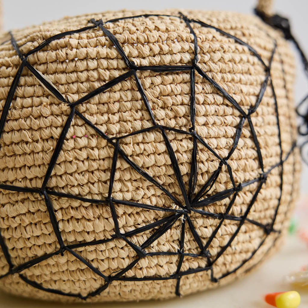 A hand-crocheted raffia Spider Basket featuring a bold black spiderweb and playful spider detail, perfectly sized for trick-or-treating, Halloween treasures, and festive fall displays.