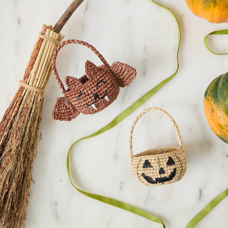 A hand-crocheted raffia Mini Bat Basket featuring playful wings, pointed ears, tiny fangs, and a sweet embroidered face for Halloween treats, treasures, and imaginative play.