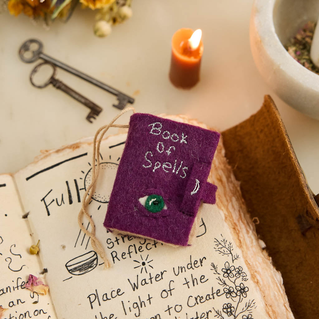 A handmade felt Book of Spells Ornament designed like a miniature mystical grimoire, complete with embroidered details, moon phases, herbal remedies, and a magical all-seeing eye.