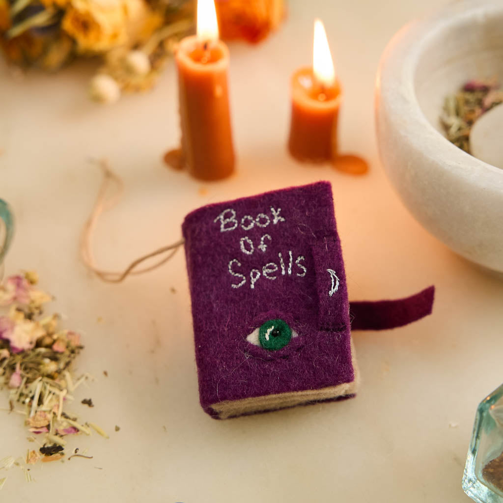 A handmade felt Book of Spells Ornament designed like a miniature mystical grimoire, complete with embroidered details, moon phases, herbal remedies, and a magical all-seeing eye.
