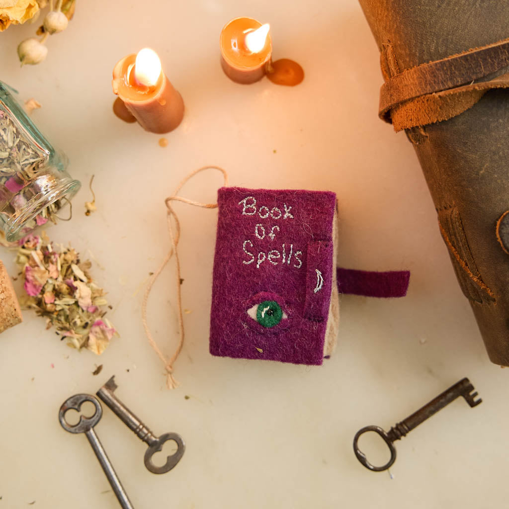 A handmade felt Book of Spells Ornament designed like a miniature mystical grimoire, complete with embroidered details, moon phases, herbal remedies, and a magical all-seeing eye.