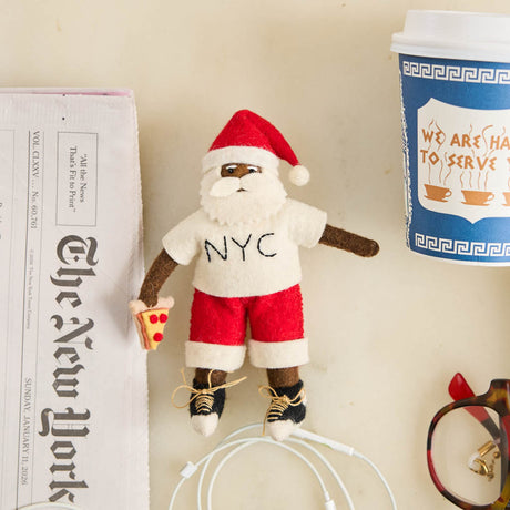 Celebrate Christmas in the Big Apple with our handmade NYC Santa Ornament, featuring a deep brown Santa dressed like a true New Yorker in an NYC tee and sneakers, with a classic slice of New York pizza in hand.
