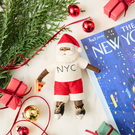 Celebrate Christmas in the Big Apple with our handmade NYC Santa Ornament, featuring a deep brown Santa dressed like a true New Yorker in an NYC tee and sneakers, with a classic slice of New York pizza in hand.