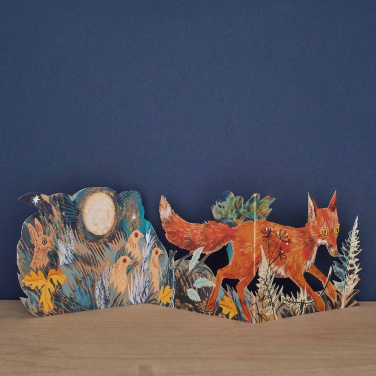Step into a moonlit woodland scene with this beautifully illustrated tri-fold card by celebrated British artist Mark Hearld. Rich in atmosphere and storytelling, the artwork captures a fox on a quiet nocturnal wander through the forest—watched closely by hens, partridge, and other woodland creatures, while a delicate bunny hides among the grasses.