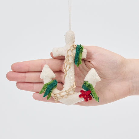 Handmade felt Anchor ornament from Craftspring held in front of a hand to show its size—featuring beaded barnacles, appliqué seaweed and starfish, and draped in netting, sustainably handmade by artisans using wool.
