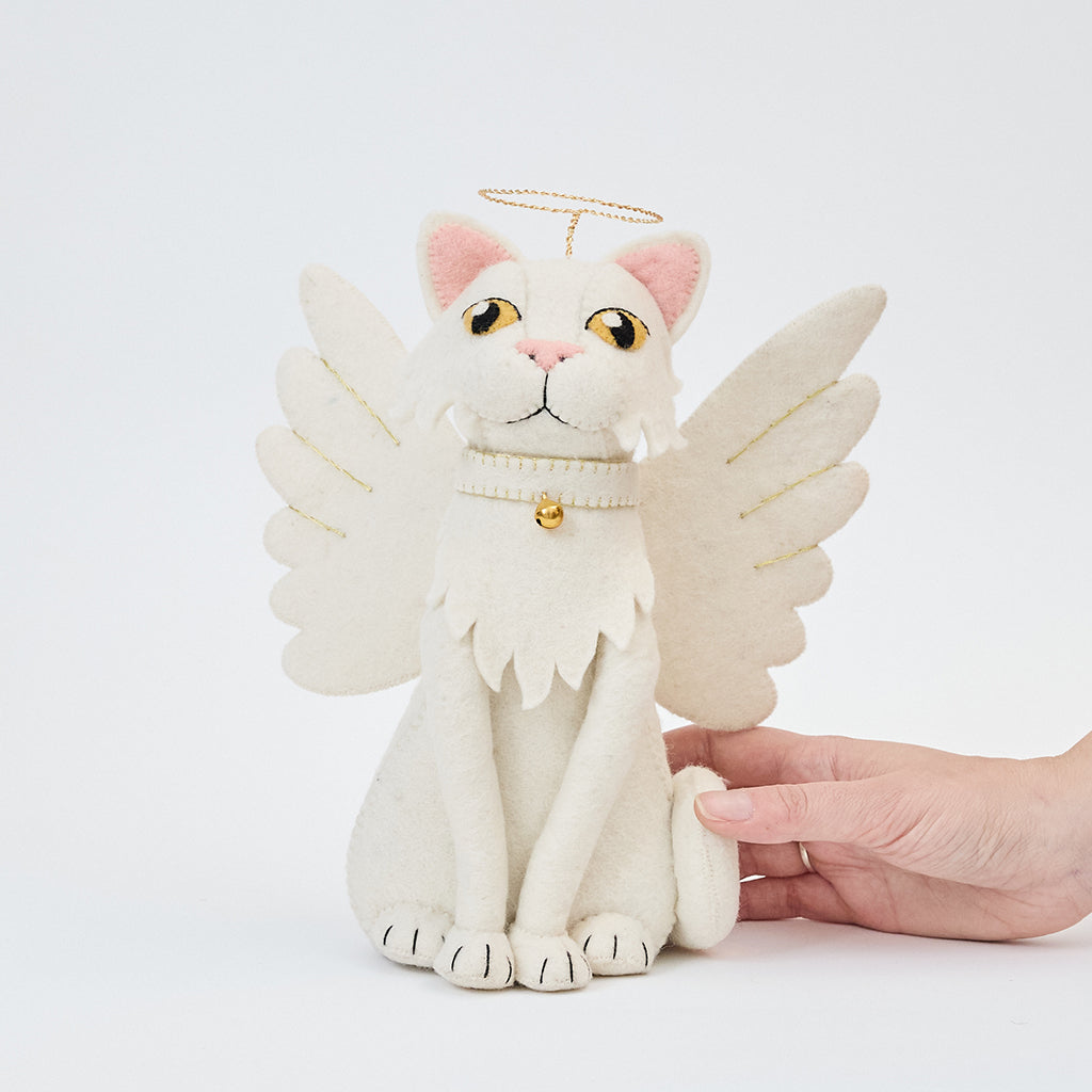 Handmade felt Angel Cat Tree Topper from Craftspring held in hand to show size—featuring white angel wings, a halo, a white collar with a bell, and gold embroidered details, ethically crafted by artisans using sustainable wool.