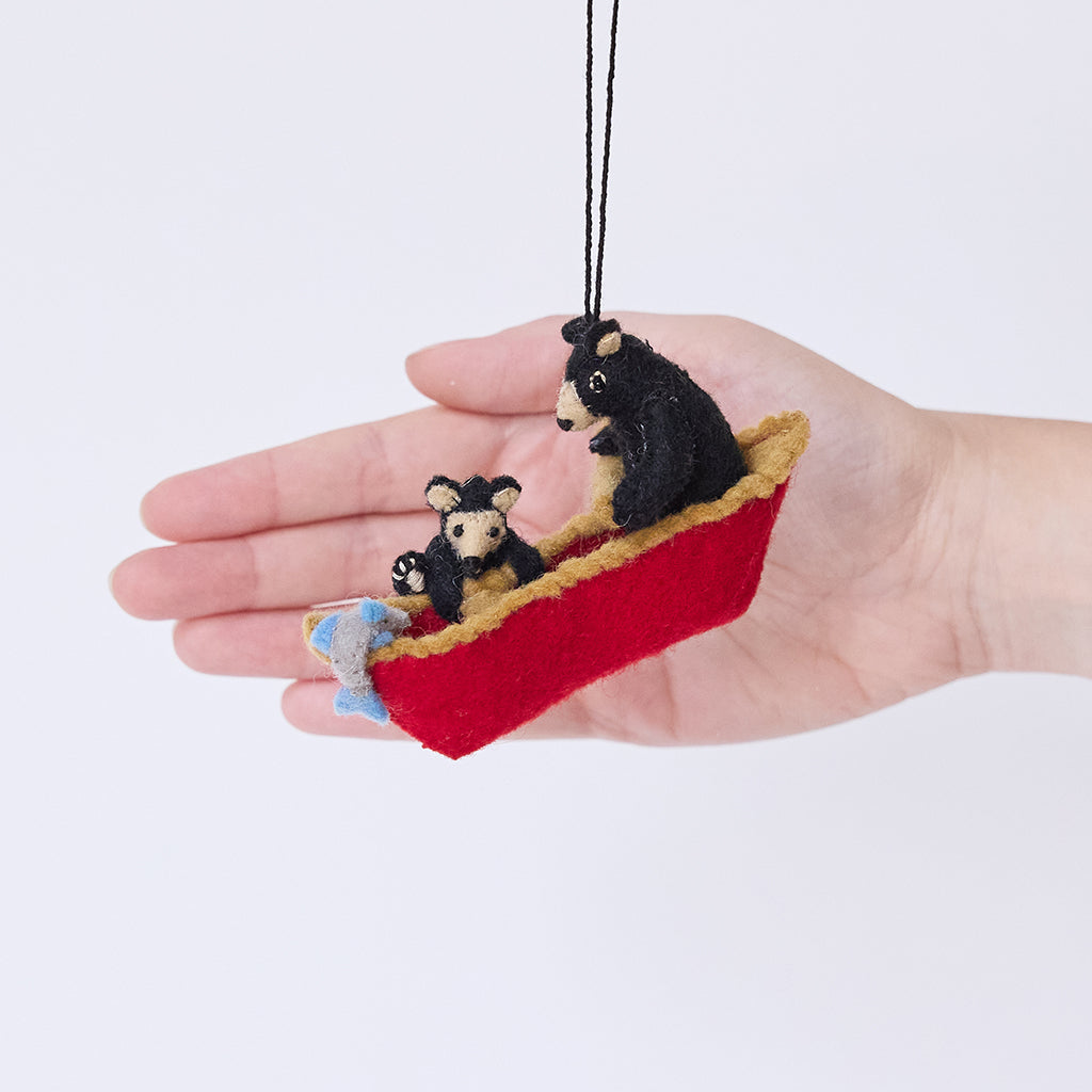 Craftspring’s Bears in a Canoe ornament held in front of a hand to show its size—handmade from sustainable wool with a mama bear paddling a canoe and a cub climbing over the side, ethically crafted by artisans using sustainable wool.