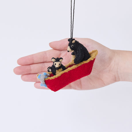 Craftspring’s Bears in a Canoe ornament held in front of a hand to show its size—handmade from sustainable wool with a mama bear paddling a canoe and a cub climbing over the side, ethically crafted by artisans using sustainable wool.